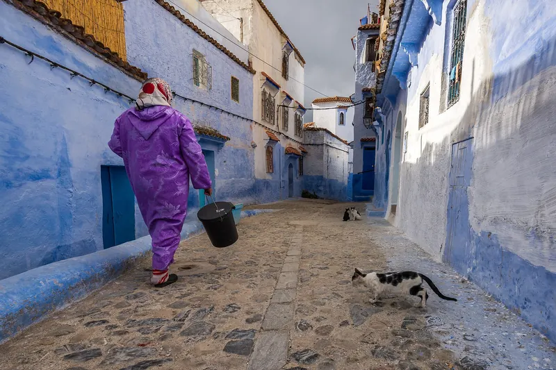 A woman in a purple djellaba walking through a blue-washed street in Chefchaouen